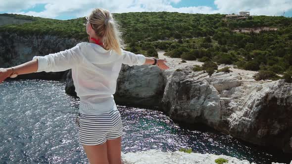 Blonde Female Tourist Wearing Shorts Standing on Cliff Spreading Arms Toward the Sea. Arms Wide Open