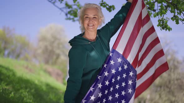 Smiling Senior Woman Holding Fluttering American Flag Looking at Camera Standing in Summer Park