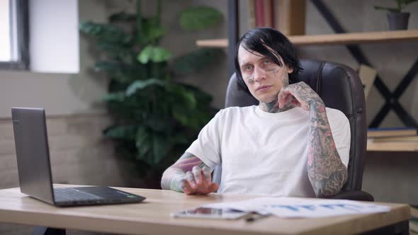 Thoughtful Tattooed Young Man Sitting at Table with Laptop Looking at Camera and Looking Around