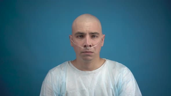 A Bald Young Man with Oncology Sadly Looks at the Camera on a Blue Background. Hair Loss Due To