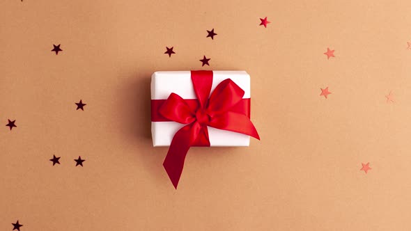 White gift box with a red satin ribbon bow tied on brown background with red stars shapes alt