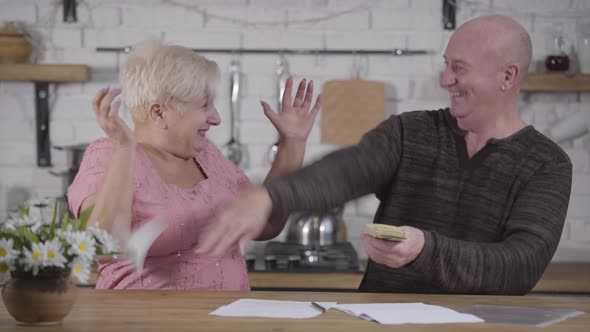 Smiling Senior Caucasian Man Showing Pack of Banknotes To His Shocked Wife. Mature Woman Throwing