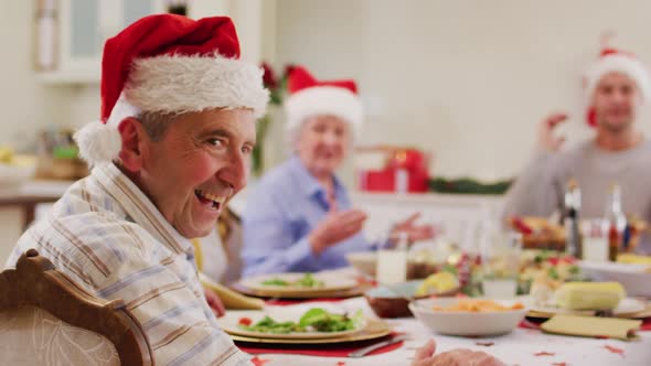 Portrait of caucasian senior man in santa hat smiling while sitting on dining table and enjoying lun