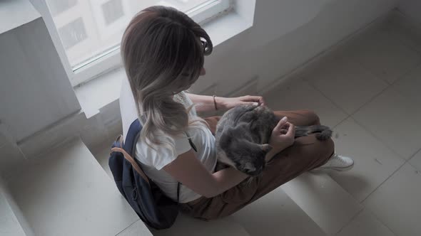 Pretty Woman Strokes Colorful Stray Cat While Sitting on White Steps of Staircase of Apartment