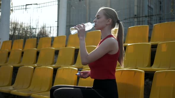 Sporty Pretty Female Sits at Staduim Tribune Drinks Water From Plastic Bottle