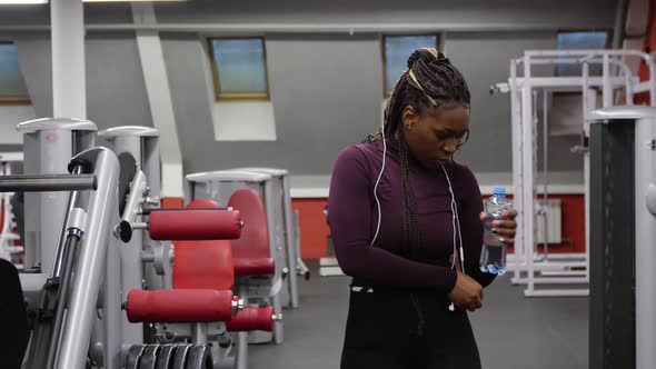 Sports Training  Africanamerican Woman Walks in the Gym and Drinking Water