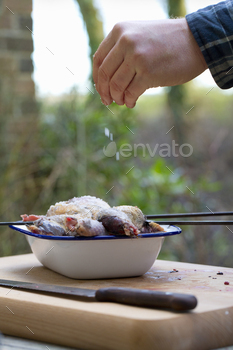 A spatchcocked game bird being seasoned for cooking, a man's hand sprinkling salt on the skin.