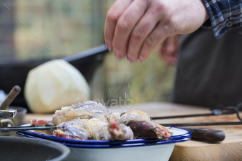 A spatchcocked game bird being seasoned for cooking, a man's hand sprinkling salt on the skin.