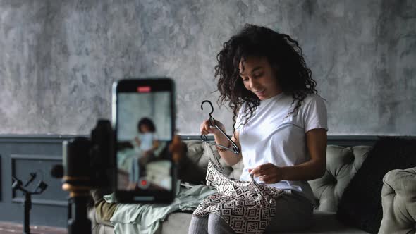 AfricanAmerican Fashion Blogger Recording Video with a Smartphone at Home Choosing Clothes Sitting