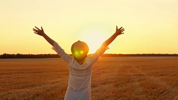 A Girl in a White Dress and a Straw Hat Raises Her Hands Up in the Wheat Field During Sunset. A