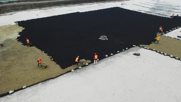 Laborers in Orange Vests Put Sand on Black Plastic Grid