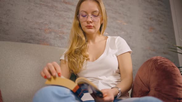 Low Angle Serious Woman Reads Book and Turns Page Sitting on Sofa