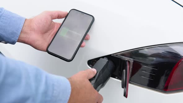 Close Up Man Inserting Power Cable Supply for Charging Electric Car and Uses the Phone