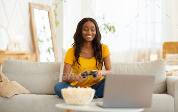 on, resting at home. Joyful African American lady gamer with joystick and popcorn sitting on couch, playing computer arcade