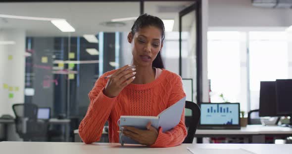 Mixed race businesswoman sitting at desk holding notebook having video call gesturing