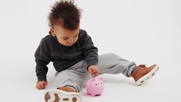 Cute AfroAmerican Toddler Sitting on the Floor and Putting Coins in the Piggy Bank Studio Shot