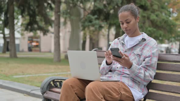 African Woman Using Smartphone and Laptop While Sitting Outdoor on Bench