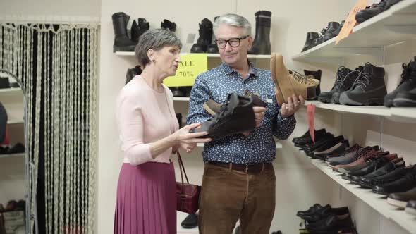 Irritated Senior Wife Putting Footwear Back on Shelves and Talking To Husband Holding Last Shoe