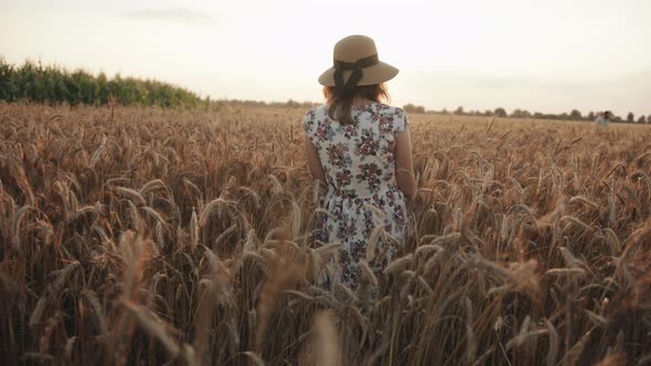 A Pleasant Young Woman in a Hat and White Summer Dress Goes Through a Wheat Field and Turns to the