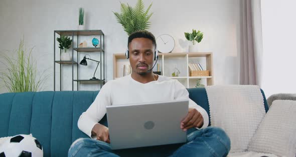 Black-Skinned Guy in Headphones Sitting on the Couch at Home and Starting to Work on Laptop