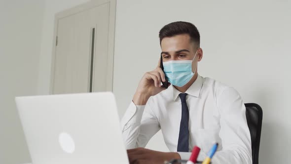 Young Man in Medical Mask Talks on a Mobile Phone When Works on a Laptop Indoors