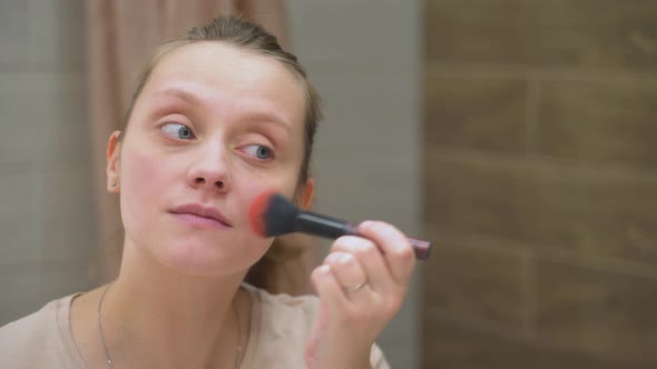 Woman with Blue Eyes is Powdering the Skin on Face with Fluffy Brush Standing By Large Mirror in the