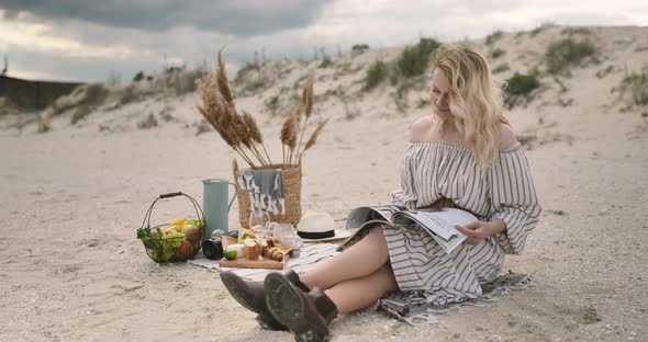 Positive model sitting on plaid on the Ocean seaside, eating fruits and read magazine.