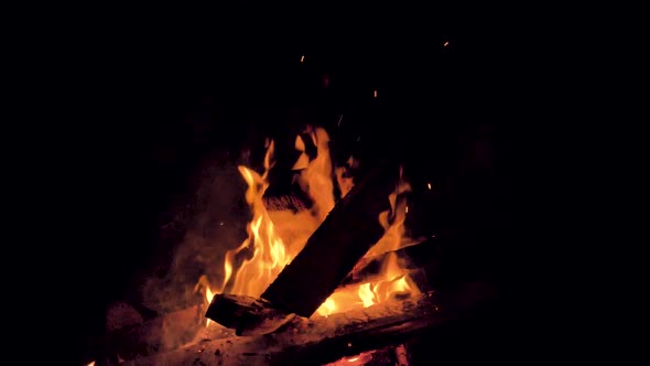 A campfire burns center frame. Darkness surrounds a glowing beach bonfire. Slow motion.