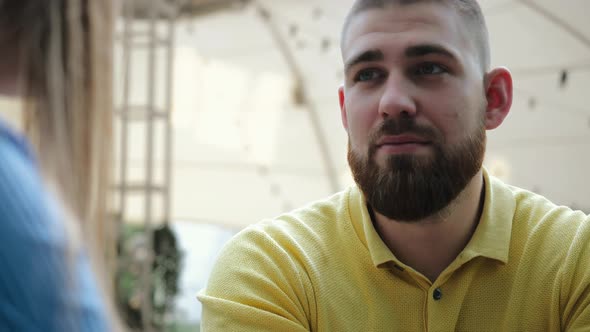 Face of Young Bearded Short-cut Guy in Yellow Polo Chatting with His Girlfriend in Outdoor Cafe