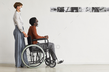 an using wheelchair and looking at paintings in modern art gallery with young woman helping him, both wearing masks, copy space