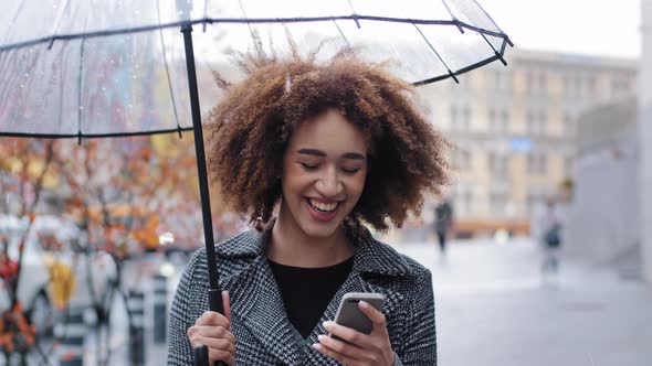 African American Girl Woman with Transparent Umbrella Stands in City in Rain Watching Funny Video on