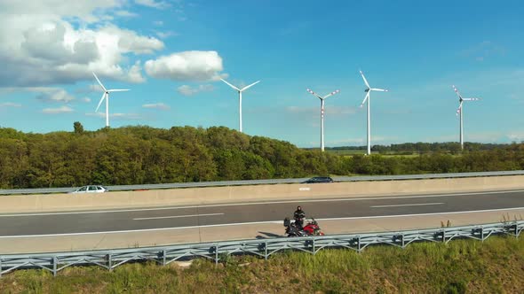 Traveler Standing on a Motorcycle on Highway, Takes Off a Drone Near Wind Farms. Austria