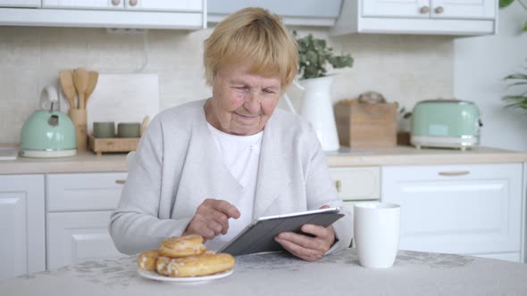 Elderly Woman Using Tablet At Home Resting On Kitchen.