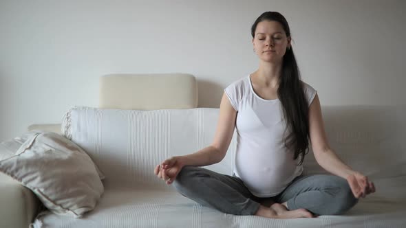 Young Pregnant Woman Sitting on White Sofa and Meditating