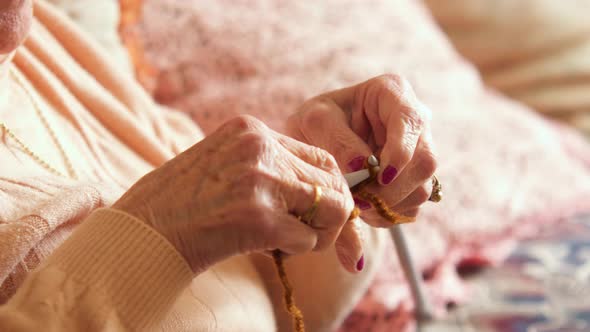 An old woman knitting in her living room, receiving sunlight, with a pink pillow behind. Her jaw is