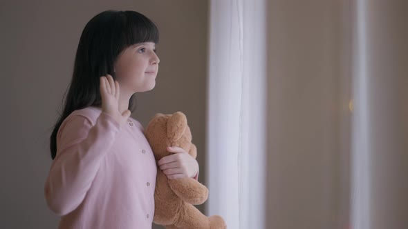 Happy Girl with Teddy Bear Looking Out Window Waving Smiling Standing in Living Room with Christmas