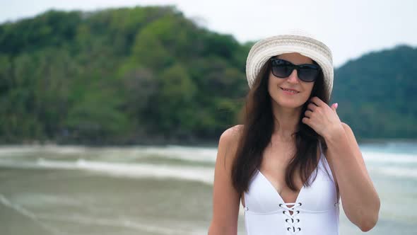 Beautiful Woman in a White Swimsuit and Hat Walking on the Beach