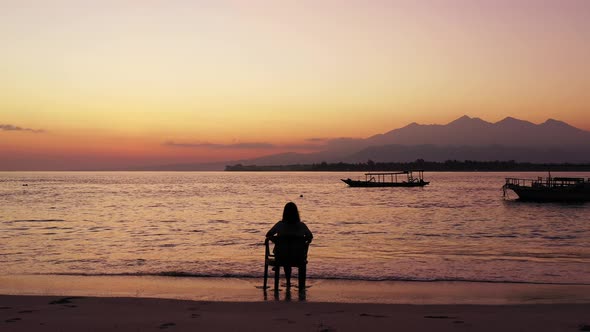 Silhouette of girl sitting on chair over sandy beach, watching colorful sky after sunset with mounta