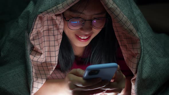 Happy Asian woman using smartphone underneath duvet in the dark on bedroom at home.