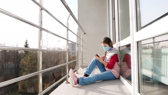 Teenager Girl in Mask and Headphones, Uses a Smartphone, Makes Selfie, on Open Balcony. Spring Sunny