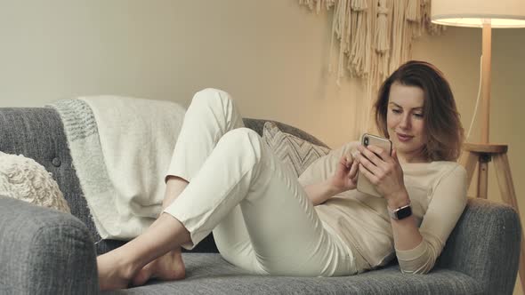 Smiling Female Scrolls Browses Phone at Home Sits on Couch at Comfy Apartment