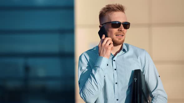 Smiling Male Businessman in Sunglasses Talking Using Smartphone Holding Briefcase Medium Shot