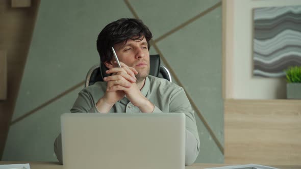 Brooding Young Man Holding Pen in Hand and Looking Away While Sitting at His Desk