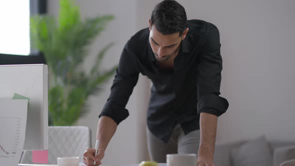 Focused Young Man Standing at Computer Table Writing Down Ideas