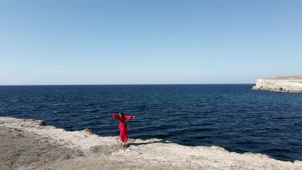 Brunette Woman in a Red Long Dress Stands on the Edge of a Cliff By the Sea