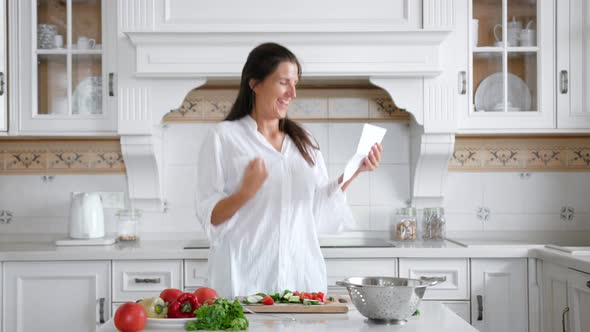 Medium Shot Pretty Young Housewife Smiling and Rejoicing Reading Letter at Kitchen While Cooking