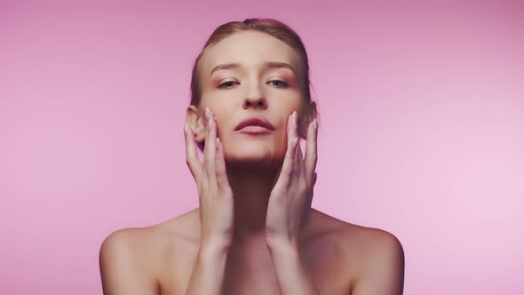 Woman with well-groomed facial skin rubs cosmetic cream with both hands on her cheeks, pink backgrou