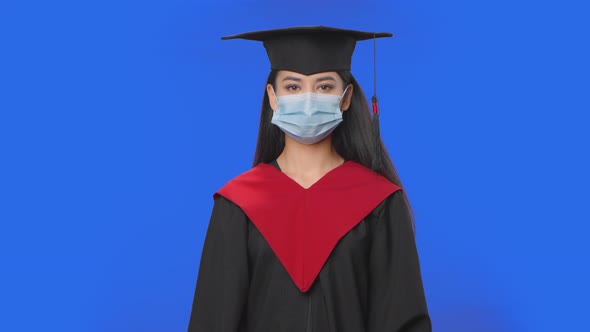 Portrait of Female Student in Cap Gown Graduation Costume and Medical Mask Looking at Camera