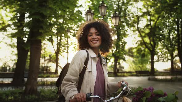 Afroamerican Female is Holding Bicycle with Lilac and Baguette in Basket By Handlebar While Posing