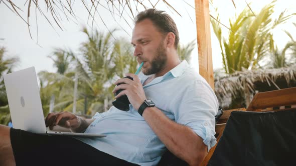 Successful Middle Aged Male Freelancer Enjoying Fruit Drink in Lounge Chair on Summer Beach